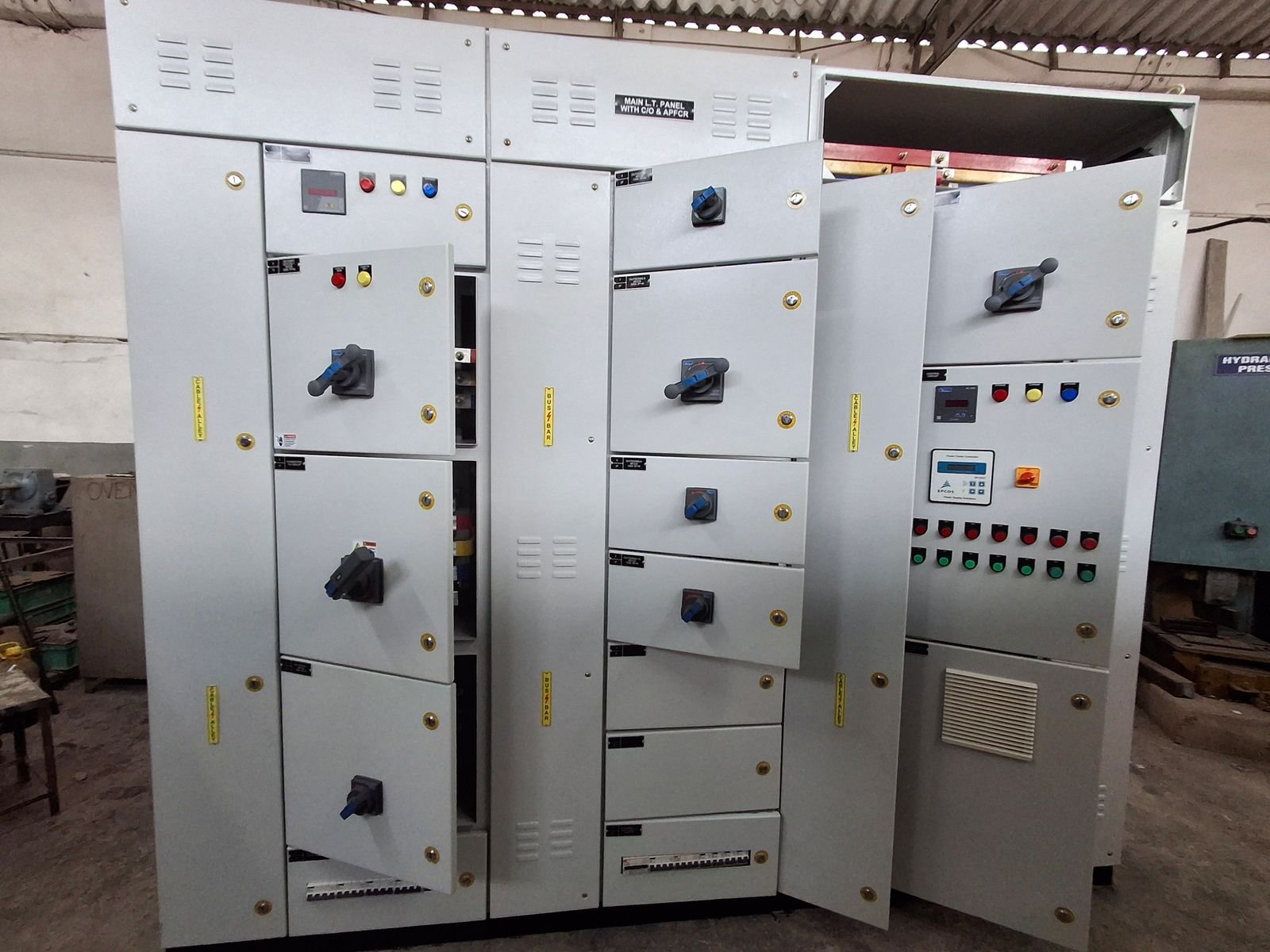 A floor-standing LT panel lineup on the works floor with several doors swung open, showing switchgear and wiring inside the compartments. The left sections carry rotary switch-disconnector handles and a digital meter; the centre section is labelled as the main LT panel with DG and power factor correction; the right-hand section carries a power factor controller above two rows of indication lamps.