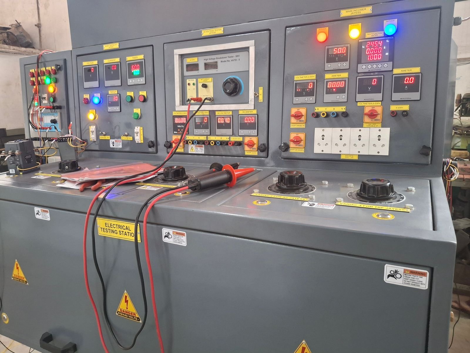 A grey testing console with four instrument panels, energised indication lamps, digital voltmeters and ammeters, four variac control knobs along the bench, and a high-voltage test lead coiled on the surface.