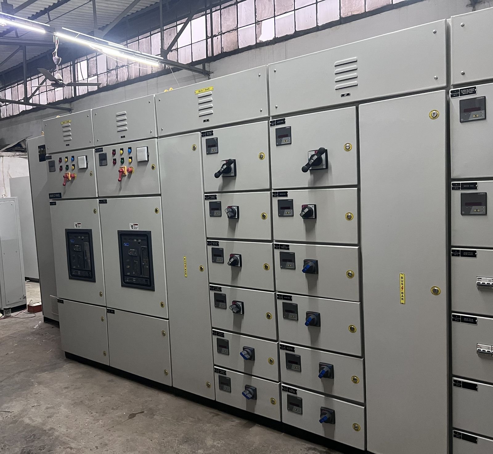 A multi-section LT panel lineup standing on the works floor seen at an angle, with two glazed breaker compartments on the near sections and columns of feeder compartments running away from the camera, under the shed roof and its fluorescent strip lighting.