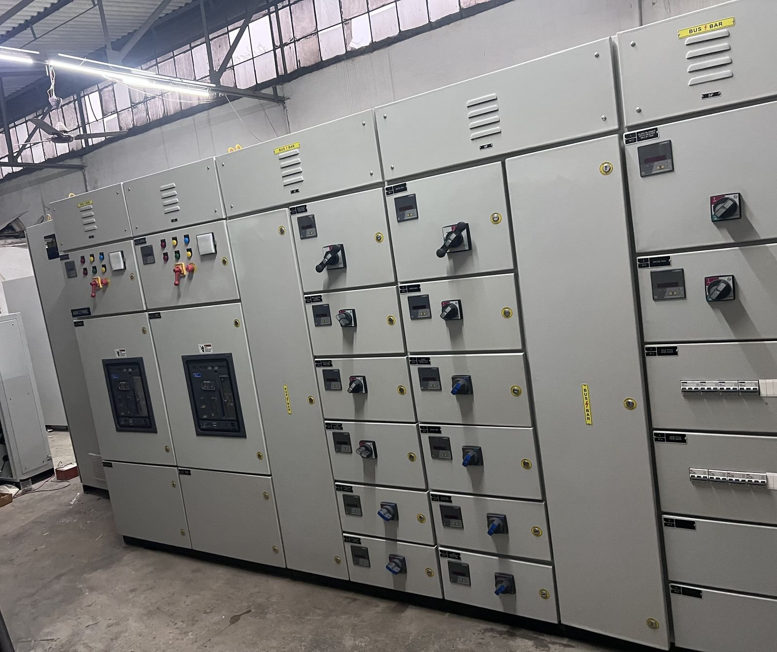 A wide view along a multi-section LT panel lineup in the works shed, the full run of compartments receding to the right, with yellow busbar chamber labels along the top edge and a second lineup part-built against the wall behind.