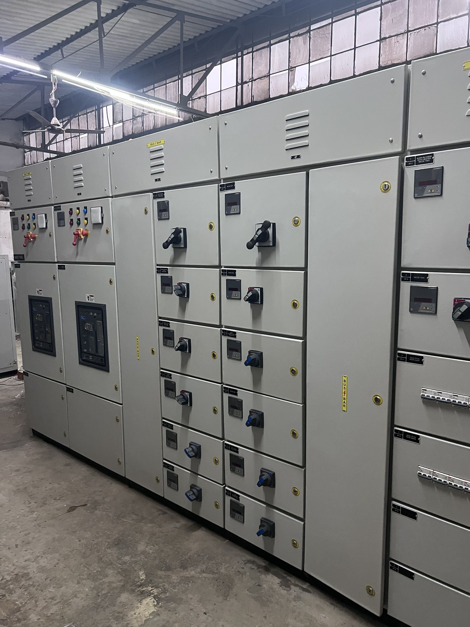 Two columns of outgoing feeder compartments in an LT panel lineup, each door carrying a digital meter, a rotary switch-disconnector handle and an engraved identification label, with glazed breaker windows on the incomer sections to the left and louvred busbar chambers above.