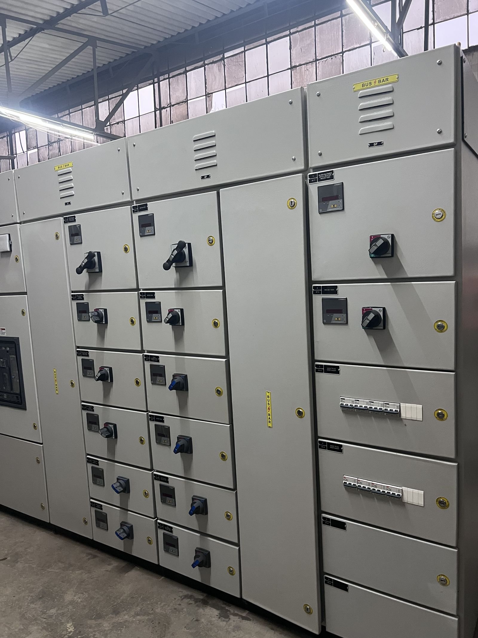 A close oblique view of an LT panel lineup showing louvred busbar chambers labelled BUS BAR along the top, columns of feeder compartments with meters and rotary handles below, and a right-hand section carrying a digital relay above two rows of miniature circuit breakers.