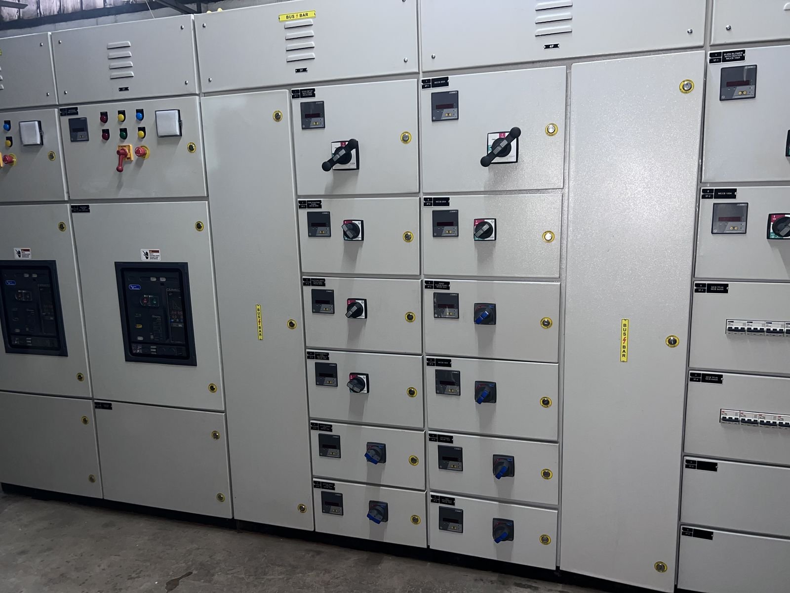 Front elevation of an LT panel lineup with two glazed incomer breaker compartments on the left, each showing the breaker facia behind the window, then columns of outgoing feeder compartments with meters and rotary switch-disconnector handles, and a right-hand section with two rows of miniature circuit breakers.