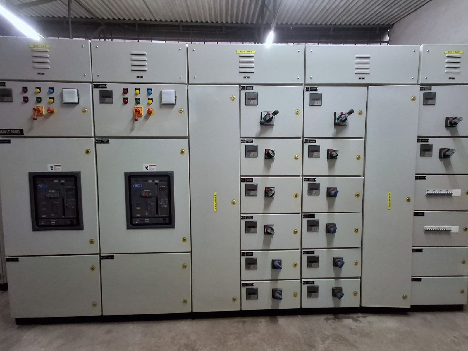 Front elevation of a multi-section low-tension panel lineup, doors closed. Two incomer sections on the left carry indication lamps, meters and rotary switch handles above glazed breaker windows; the sections to the right are columns of outgoing feeder compartments, each with a meter and a rotary switch-disconnector handle, with louvred busbar chambers along the top.