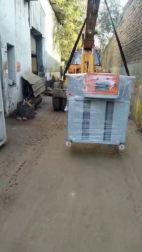 A shrink-wrapped floor-standing panel with an orange-fronted control section on top, slung from a hydraulic crane and moving down a walled lane away from the works.