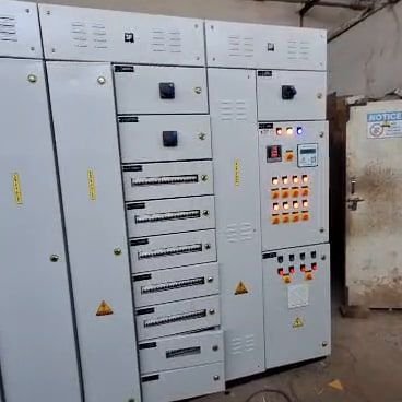 A multi-section LT panel lineup indoors under a metal roof, indicator lamps lit across several compartments, rotary switches and ventilation louvres on the doors, with a pedestal fan alongside.