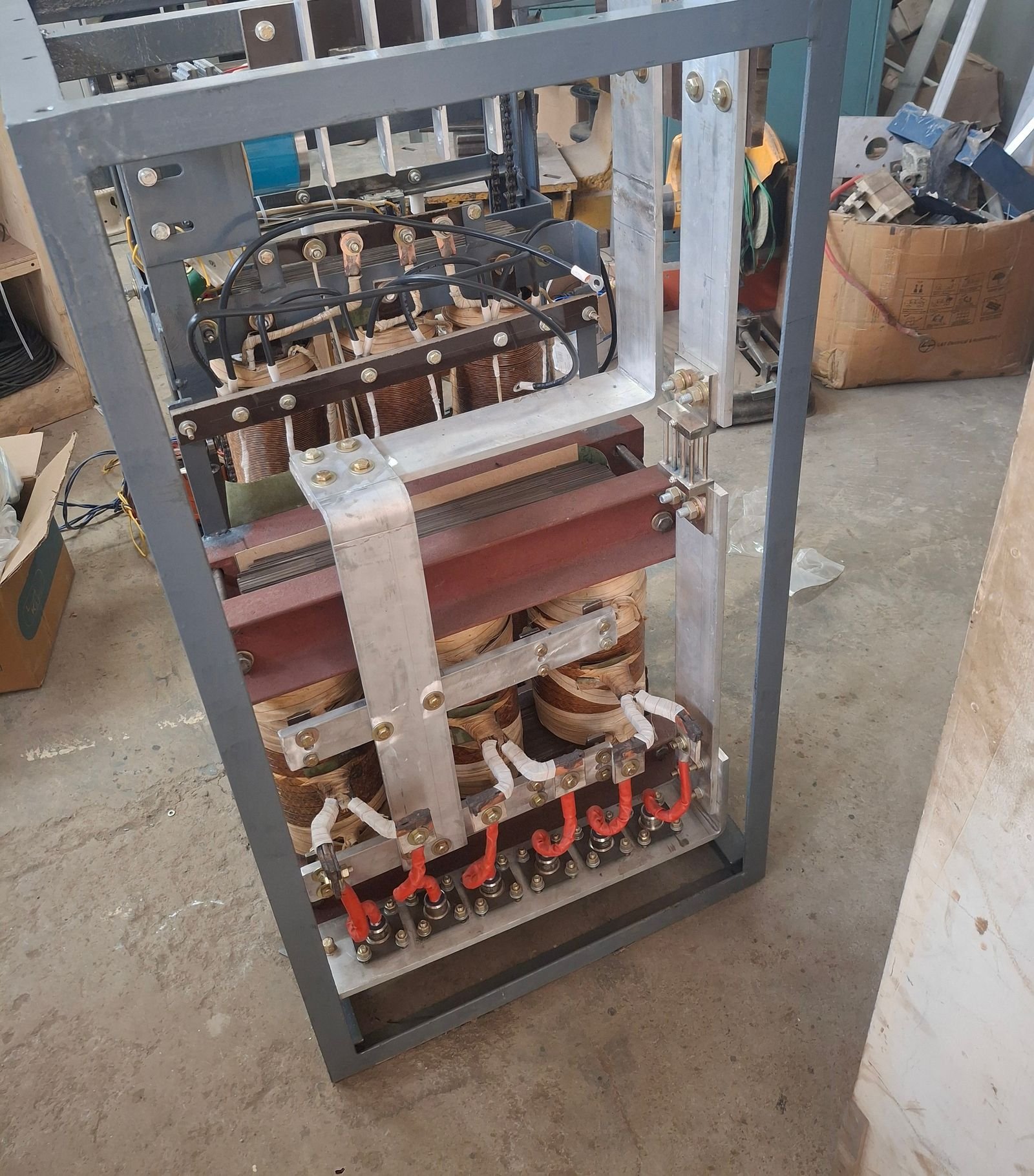 A servo voltage stabiliser part-assembled in its steel frame: strip-wound buck-boost coils on a laminated core above, variable winding assemblies below, joined by aluminium busbar links to an insulated terminal board at the base.