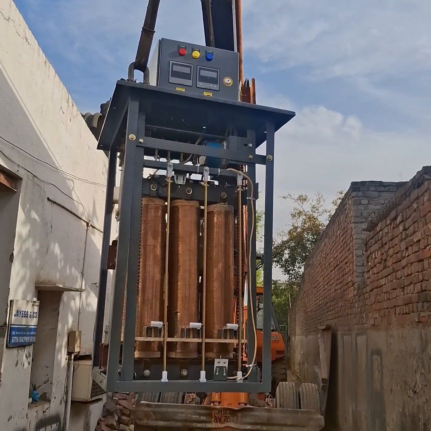 A three-phase transformer core-and-coil assembly on a steel frame, copper windings visible on all three limbs, suspended from a hydraulic crane in a works yard.